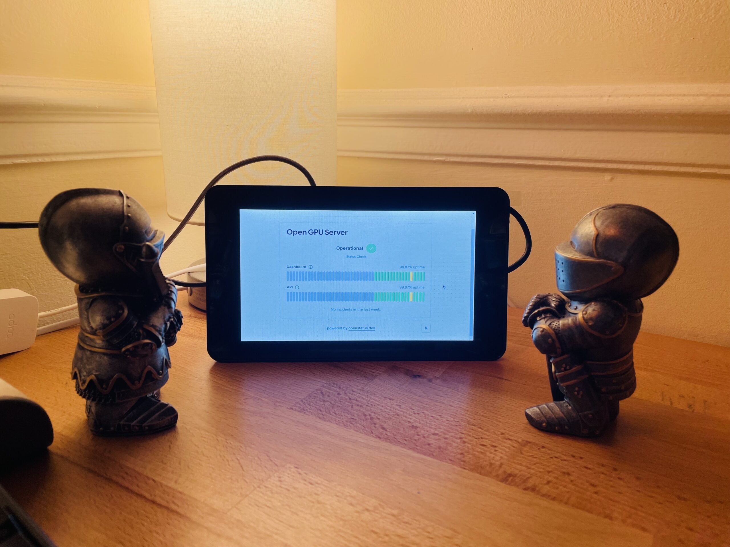 A small tablet or display screen sits on a wooden surface, showing the interface for an "OpenGPU Server" with status bars indicating it's operational. On either side of the screen are two small figurines of armored guards. The scene is set against the background of a wall with decorative molding, giving it a home-like setting.